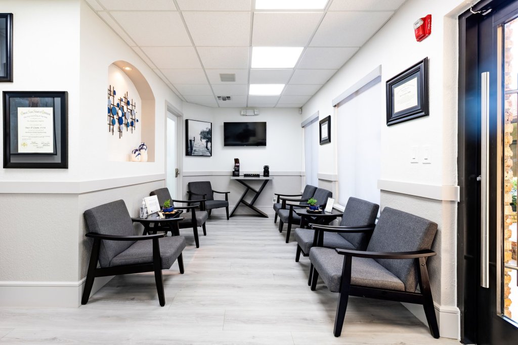 Modern waiting room with rows of black armchairs, white walls with framed artwork, wall-mounted television, and light wood-style flooring