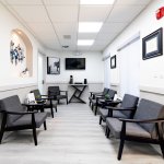 Modern waiting room with rows of black armchairs, white walls with framed artwork, wall-mounted television, and light wood-style flooring