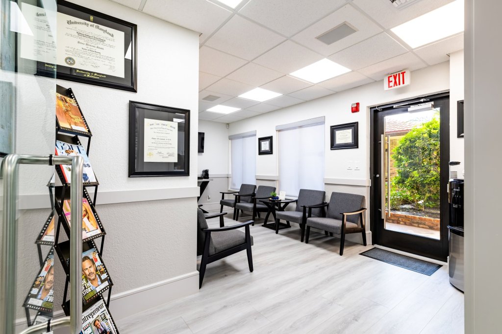Waiting area view showing magazine rack, black seating, framed certificates on wall, and glass exit door with view of outdoor greenery