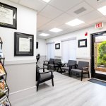 Waiting area view showing magazine rack, black seating, framed certificates on wall, and glass exit door with view of outdoor greenery