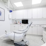 Modern white dental operatory with patient chair, white cabinetry, wall-mounted artwork, and clinical equipment in bright sterile environment