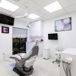 Contemporary dental treatment room with grey patient chair, multiple wall-mounted monitors, windows with outdoor views, and skylight illumination