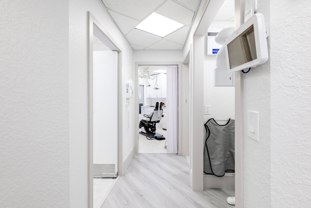 Bright hallway with skylight illumination, wall-mounted monitors, view into treatment operatory with dental chair, and light professional finishes