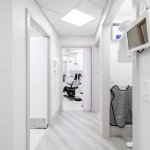 Bright hallway with skylight illumination, wall-mounted monitors, view into treatment operatory with dental chair, and light professional finishes
