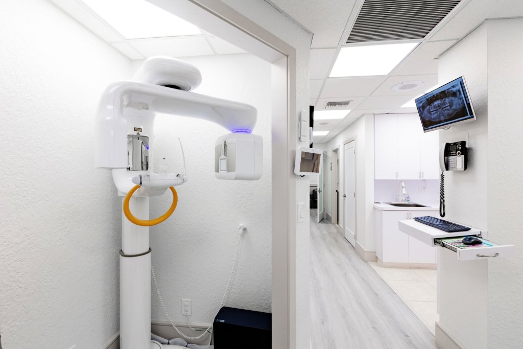 Hallway showing dental panoramic X-ray machine with yellow accents, wall-mounted monitors, and view into treatment areas with modern white design