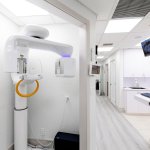 Hallway showing dental panoramic X-ray machine with yellow accents, wall-mounted monitors, and view into treatment areas with modern white design