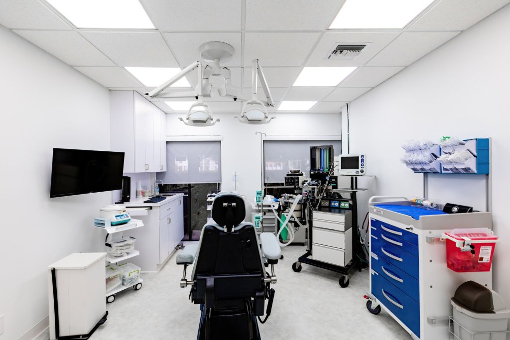 Medical treatment room with black patient chair, blue instrument cart, multiple mobile equipment carts, wall-mounted monitor, and clinical supplies
