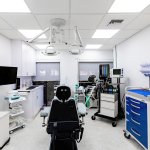 Medical treatment room with black patient chair, blue instrument cart, multiple mobile equipment carts, wall-mounted monitor, and clinical supplies