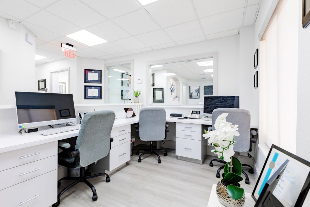 Modern administrative office area with white desks, grey office chairs, computer workstations, white cabinetry, and decorative white orchid plant