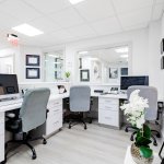 Modern administrative office area with white desks, grey office chairs, computer workstations, white cabinetry, and decorative white orchid plant