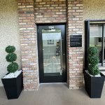 Entrance to dental office featuring brick wall, glass door with black frame, and two black planters with manicured topiary trees