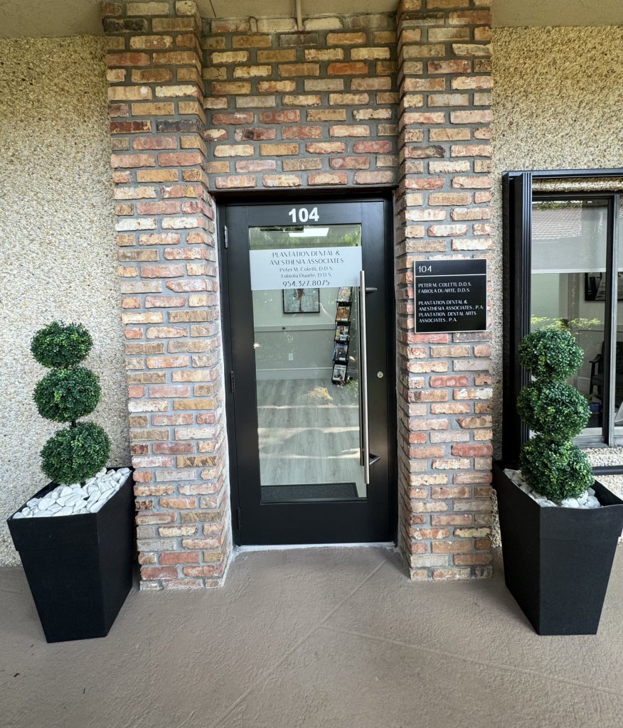Entrance to dental office featuring brick wall, glass door with black frame, and two black planters with manicured topiary trees